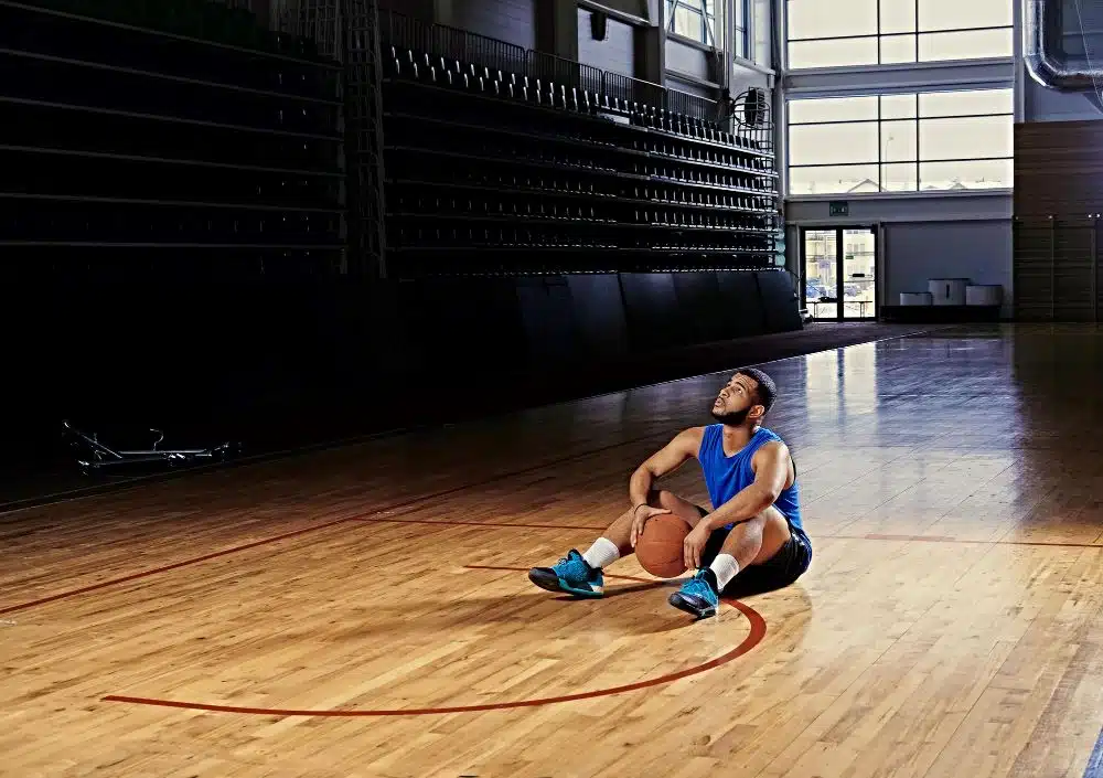 A professional basketball player sits on the floor of a basketball court looking up to the sky holding a basketball in his hands